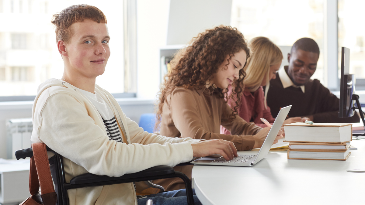 Young man in a wheelchair at a shared desk with a laptop, studying alongside other students.