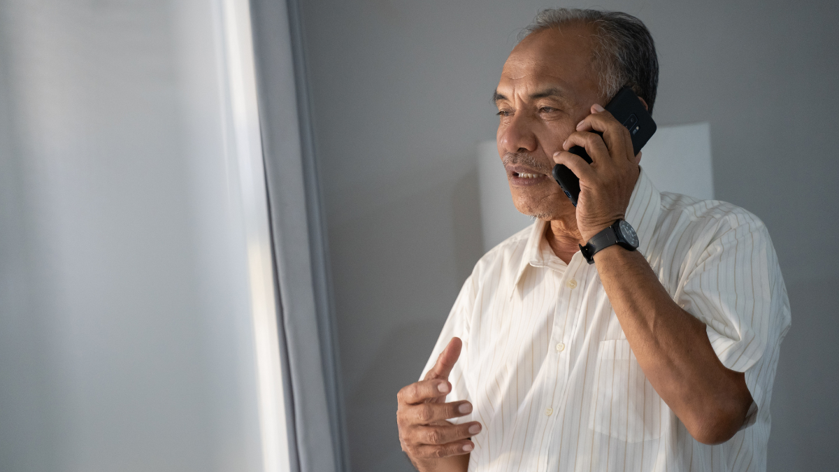 Older man talking on a mobile phone beside a window, as aged care clients and families do when they enquire.